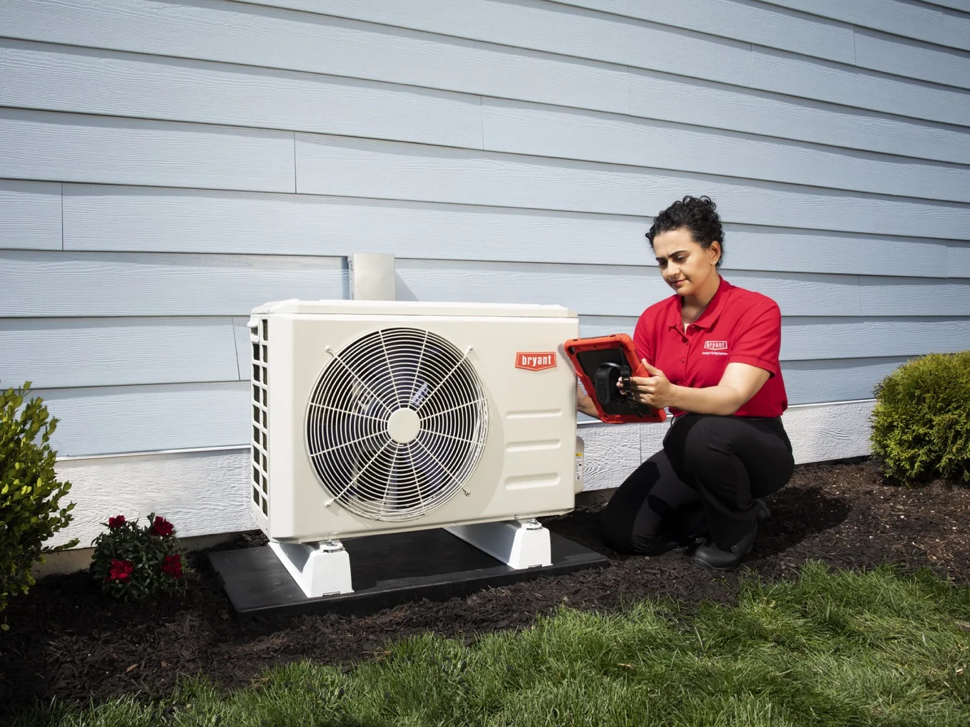 A Bryant technician at a ductless mini-split outdoor unit, checking the system on a tablet